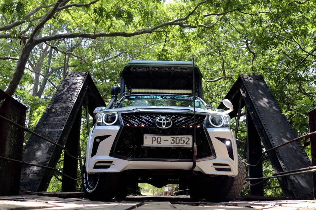 White SUV parked on an iron bridge, viewed from below. Sunlight filters through lush green trees above, casting dappled shadows on the vehicle.