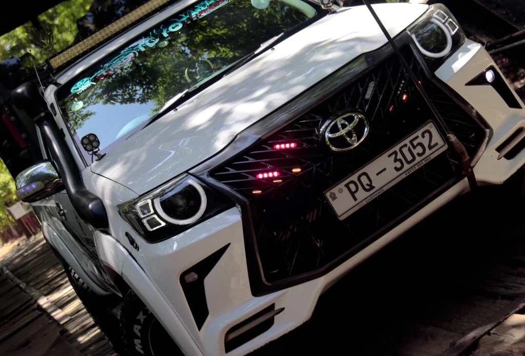 Custom white Toyota truck with neon lights and off-road gear, parked on a shaded path. The front grill features a bold design, creating an adventurous vibe.