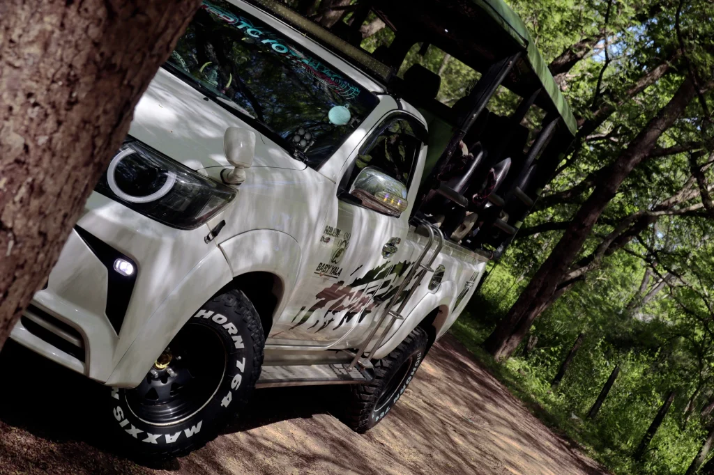 A modified white safari truck with bold tires is parked on a dirt path in a lush green forest, conveying a sense of adventure and exploration.