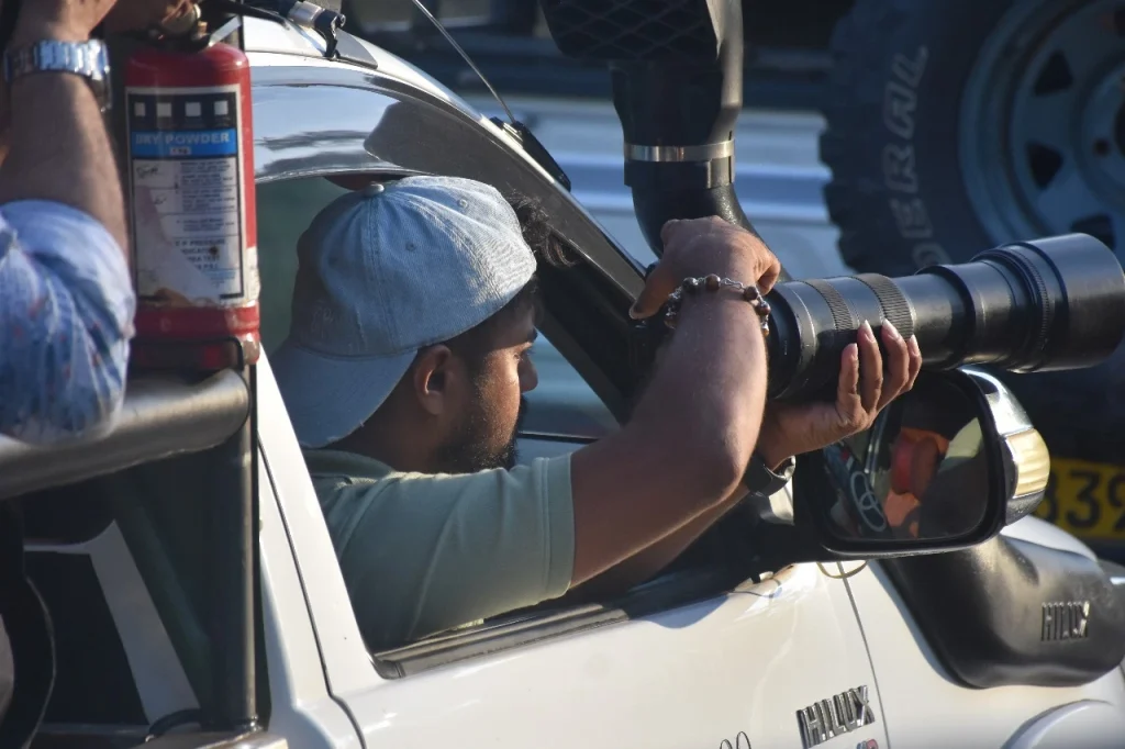 Man with a cap uses a large telephoto lens to take photos from a white vehicle's open window. There's a fire extinguisher mounted nearby.